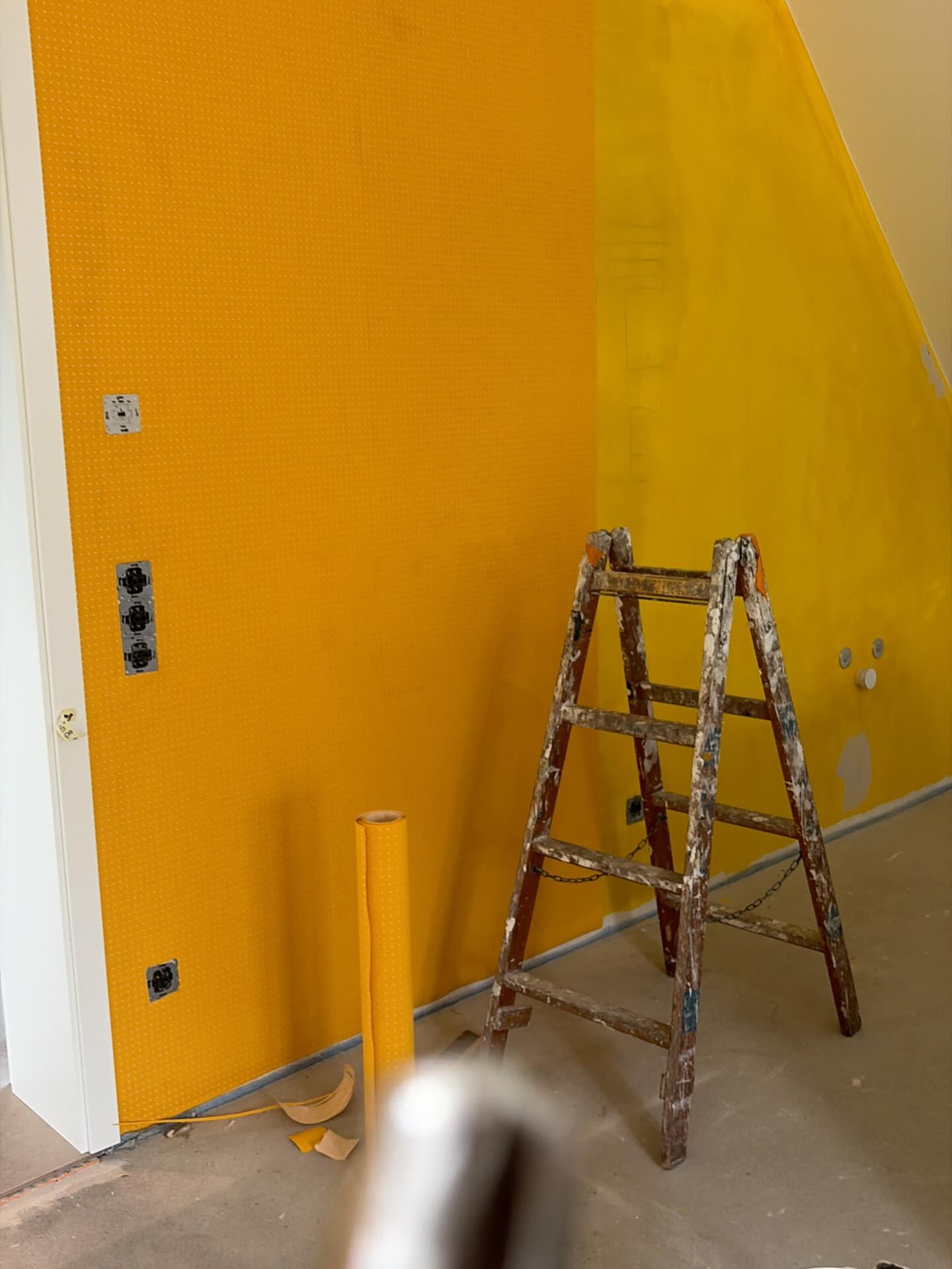 Gelb gestrichene Wand mit einer Holzleiter, am unteren Bildrand eine Farbrolle.