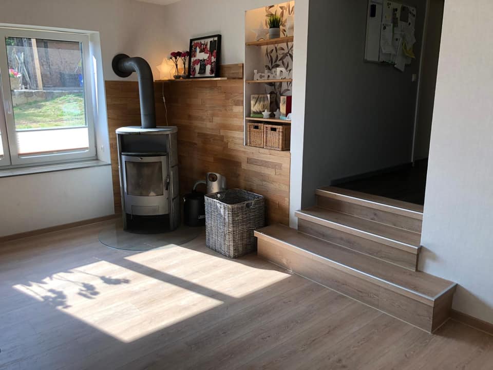 Heller Wohnraum nach der Renovierung mit Kaminofen vor einer Holzwand, Stufen und Tageslicht.