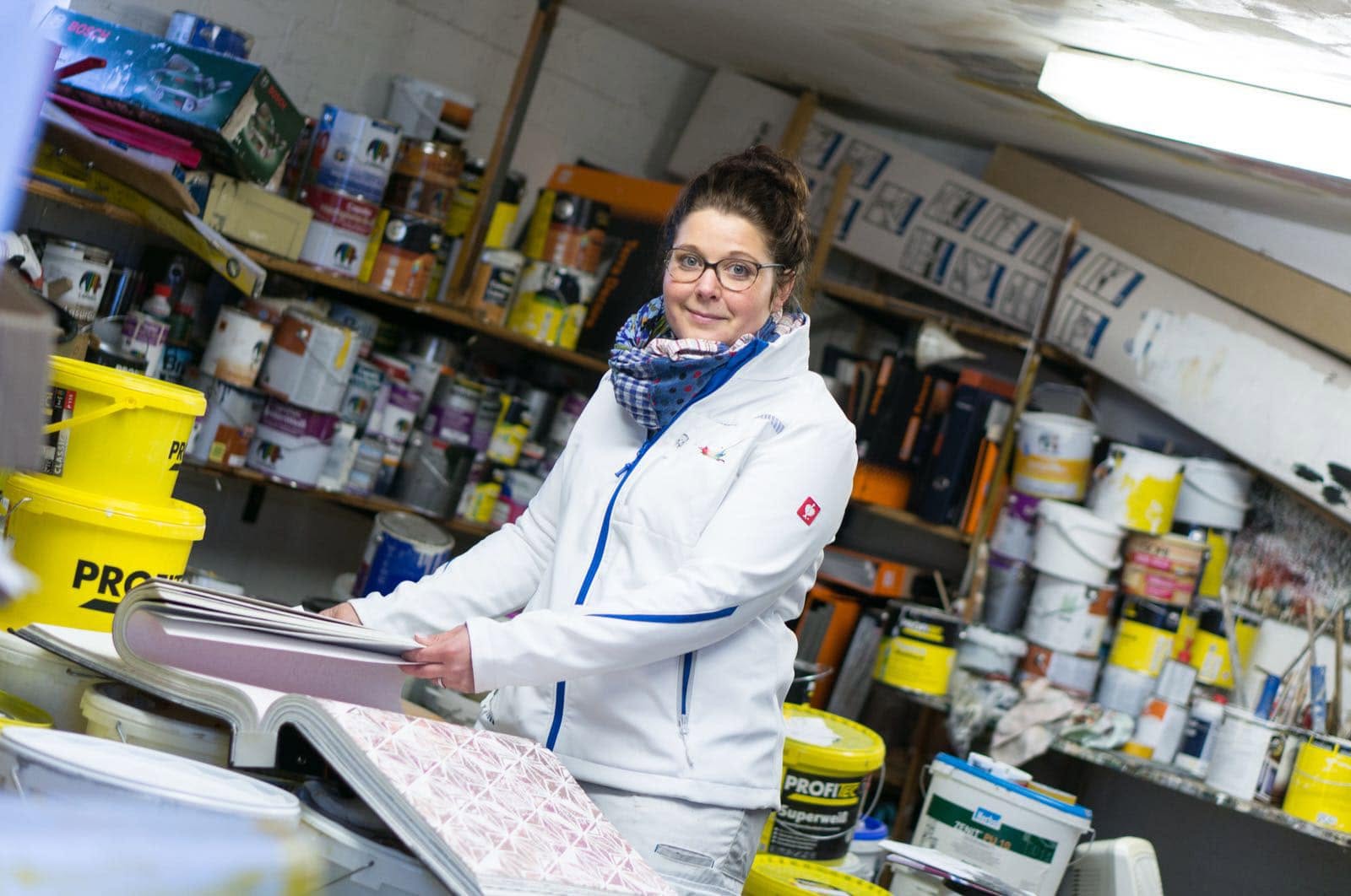 Lisa Louis in weißer Arbeitsjacke am Farbmischplatz vor Regalen voller Farbeimer, sie lächelt in die Kamera.