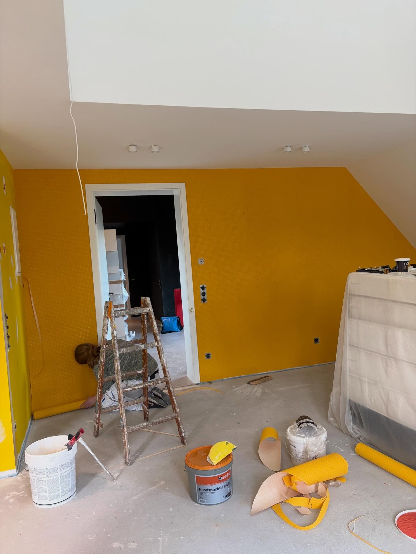 Raum unter der Dachschräge während der Arbeit: gelb gestrichene Wand, davor eine Holzleiter, Eimer und Tapetenrollen auf dem Boden.