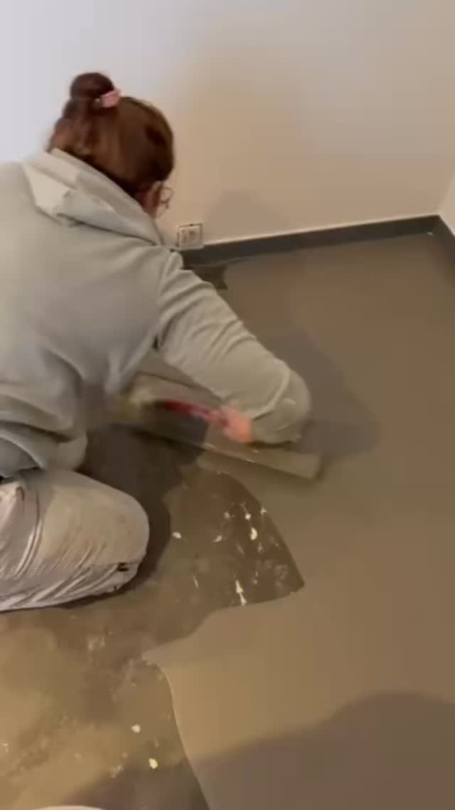 Lisa Louis kniet auf dem Estrich und verteilt Ausgleichsmasse mit der Glättkelle.