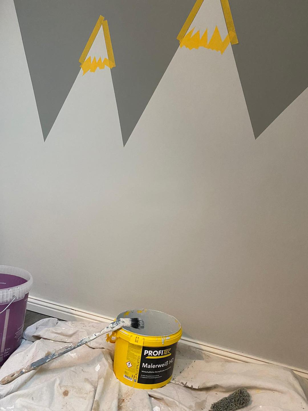 Bergmotiv in Arbeit, die Gipfel mit gelbem Klebeband abgeklebt, am Boden Abdeckvlies und Farbeimer.